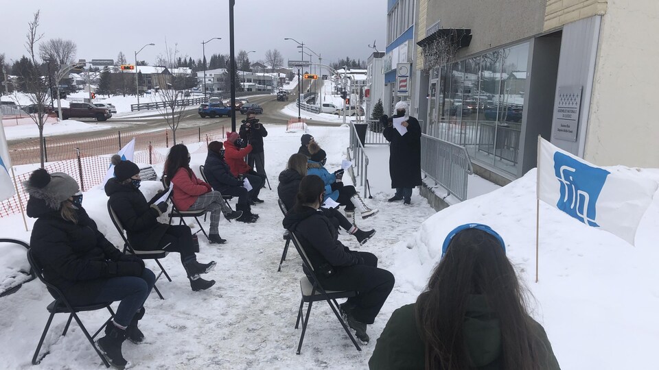 Une dizaine de personnes sont assises sur des chaises pliantes placées sur le trottoir devant le bureau de la députée Suzanne Blais. Tous les membres portent un couvre-visage.