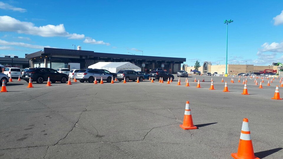 Des voitures en file, avec des cônes pour délimiter les couloirs, devant un immeuble sur un terrain de stationnement d'un centre commercial.