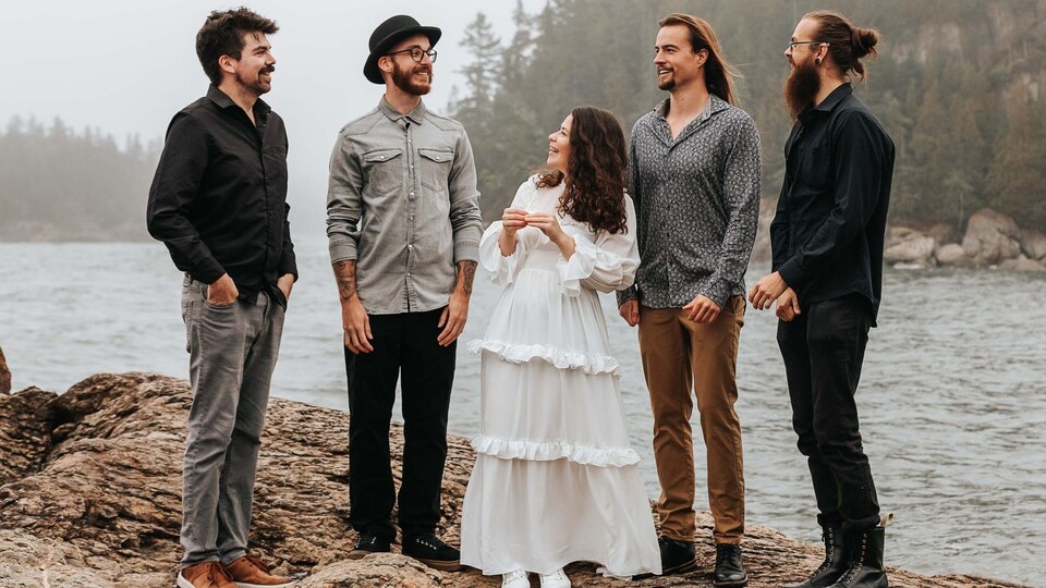Émilie Lévesque et son groupe musical Fleurs, composé de quatre musiciens. Ils sont sur le bord de l'eau et se sourient mutuellement. Elle porte une robe.