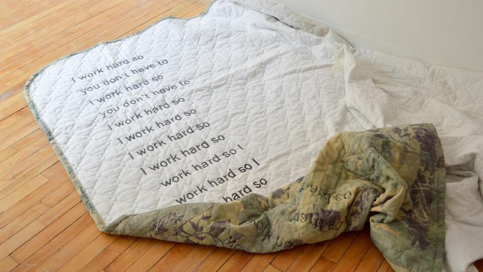Une couverture sur un plancher, sur laquelle sont brodé les mots «I work hard so you dont have to».
