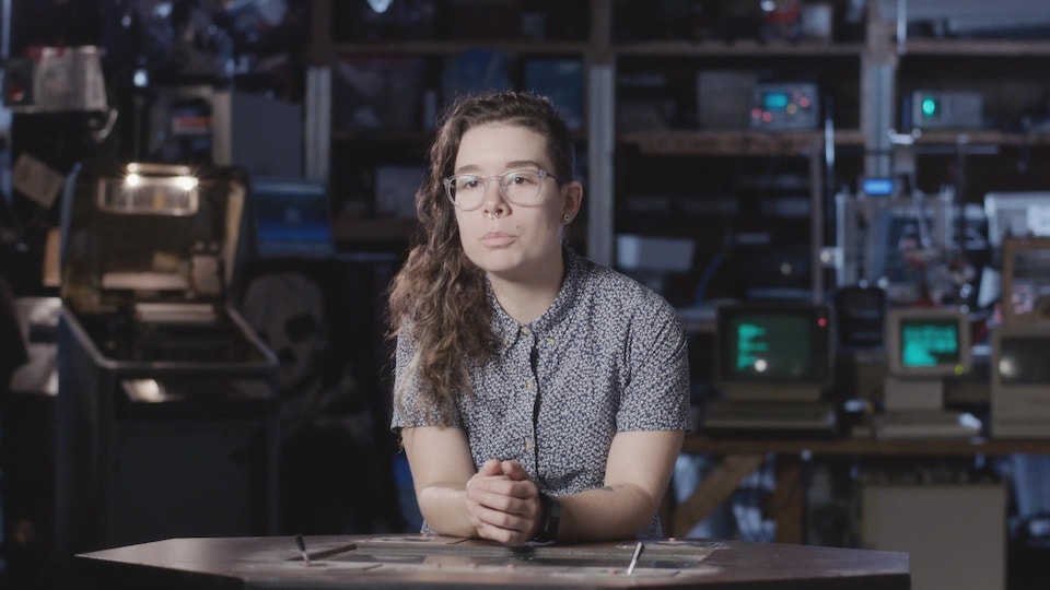 Une jeune femme aux cheveux longs bouclés accoudée à un bureau, en train de parler à quelqu'un hors du cadre. Elle est assise devant un bric à brac de vieux matériel informatique.