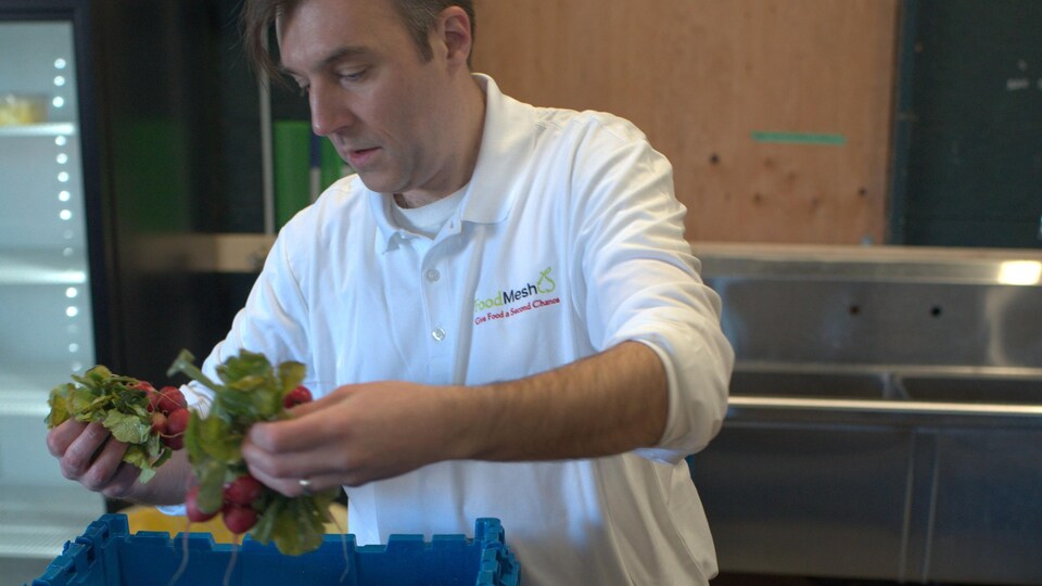 Un homme s'apprête à déposer des radis avec leur feuillage dans un contenant en plastique bleu. Dans une boîte de carton brune à côté du contenant bleu, il y a des carottes, des poivrons, des oignons et des radis.