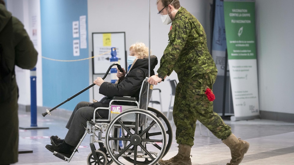 Un membre des Forces armées canadiennes transporte une dame en fauteuil roulant.