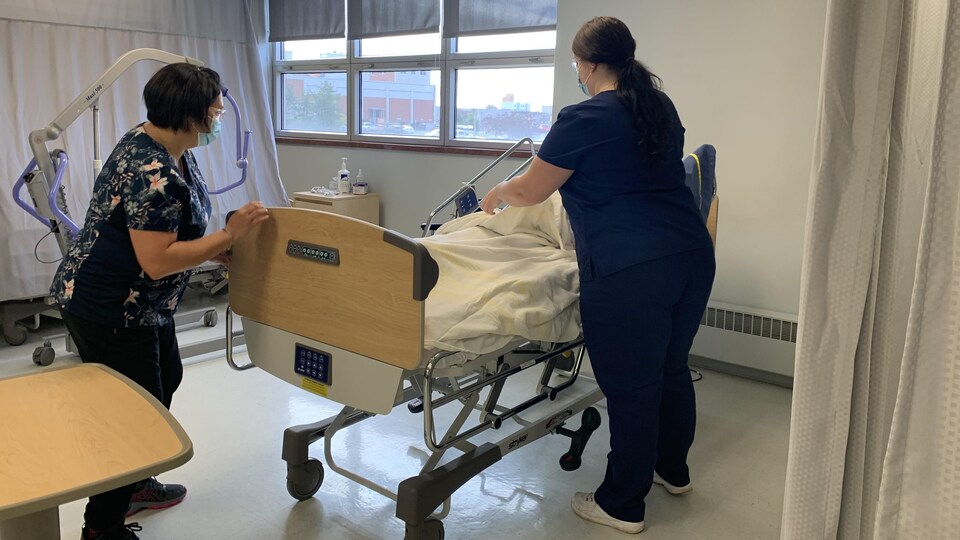 Deux femmes manipulent un lit d'hôpital où est couché un mannequin.