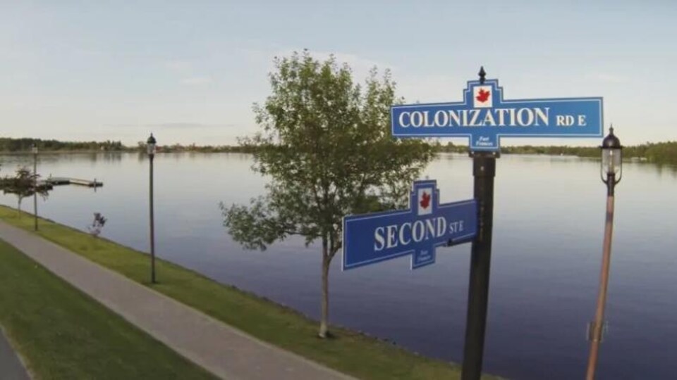 Sur le bord d'une rivière, un chemin avec une pancarte où on voit le nom des rues Colonisation et Second street.