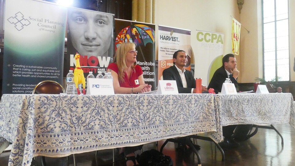 Trois candidats assis à une table, devant une série de panneaux promotionnels d'organismes communautaires, dans une grande salle.