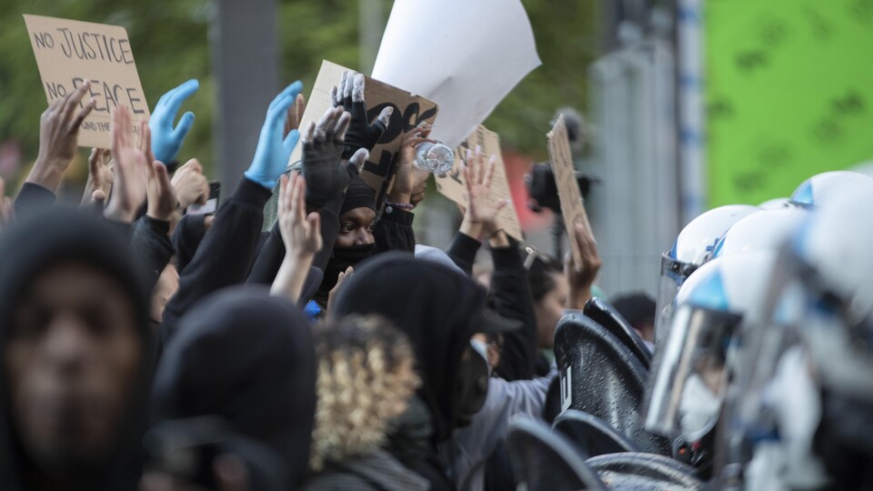 Un groupe de manifestants, la main levée, certains portant des pancartes, font face aux policiers portant le casque.