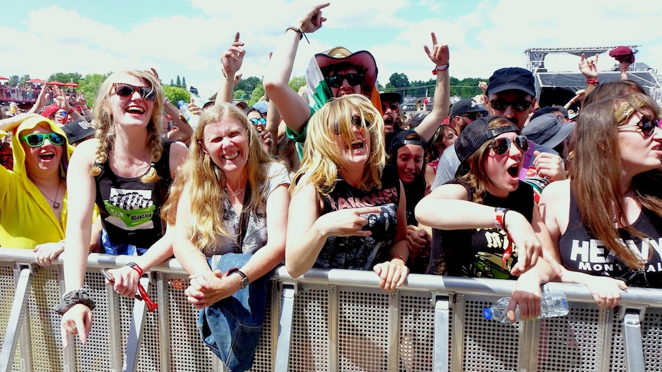 Des femmes souriantes et enthousiastes sont alignées le long d'une clôture lors d'un spectacle du Rockfest en plein soleil.