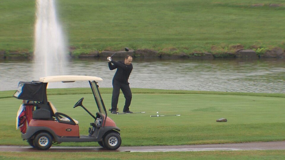 Un golfeur prend son élan pour frapper une balle sur le bord d'un étang d'où jaillit un jet d'eau.
