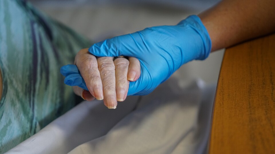 Une main ganté tient dans sa main une main d'une personne âgé couché dans un lit.