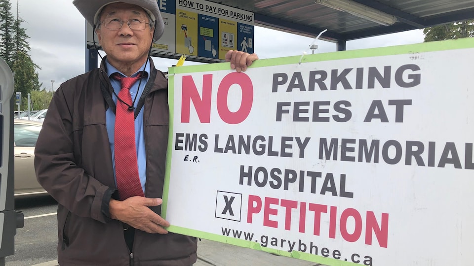 L'homme portant un manteau et un chapeau brandit une pancarte devant les parcomètres de l'hôpital.