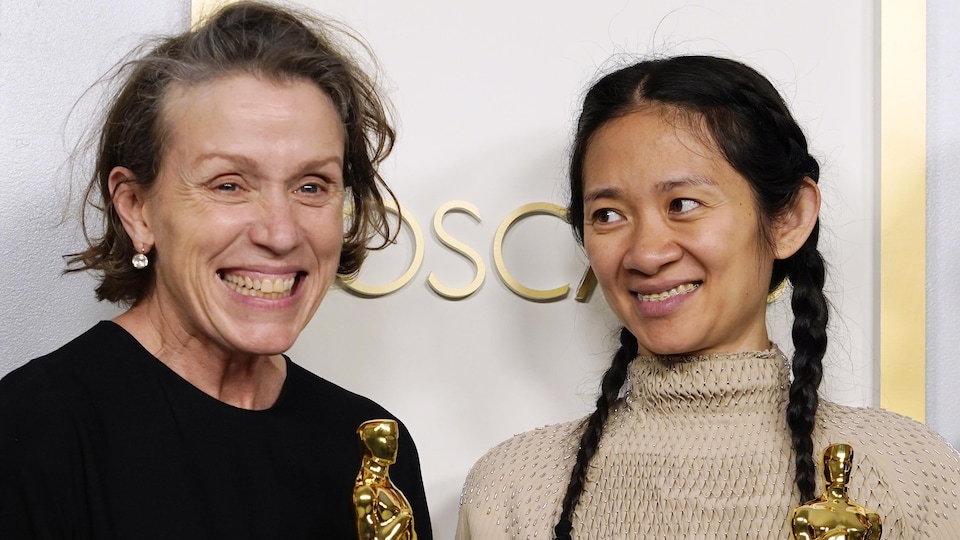 Deux femmes sourient, chacune tient un trophée dans ses mains. Elles sont debout devant un mur où l'on devine l'inscription OSCAR en lettres or.