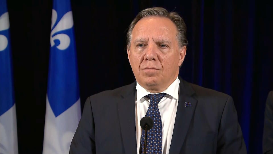 François Legault, debout devant un lutrin, parle devant un micro. Il porte un veston et une cravate. Des drapeaux du Québec en arrière-plan.