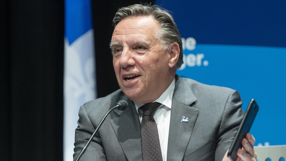 François Legault tenant un téléphone dans sa main gauche lors d'une conférence de presse.