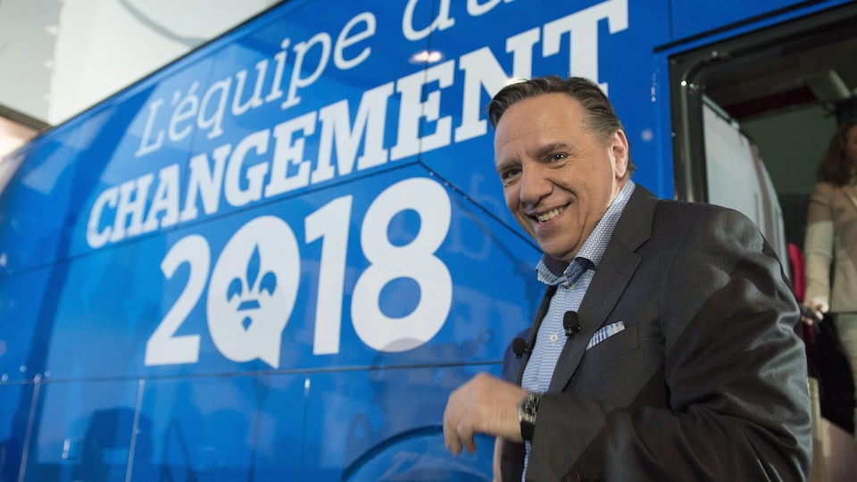 François Legault sourit pour les caméras alors qu'il se trouve devant l'autobus de la CAQ, sur laquelle est inscrit « L'équipe du changement 2018 ». 