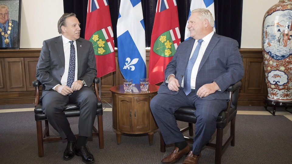 Deux hommes, assis devant des drapeaux de l'Ontario et du Québec, discutent.