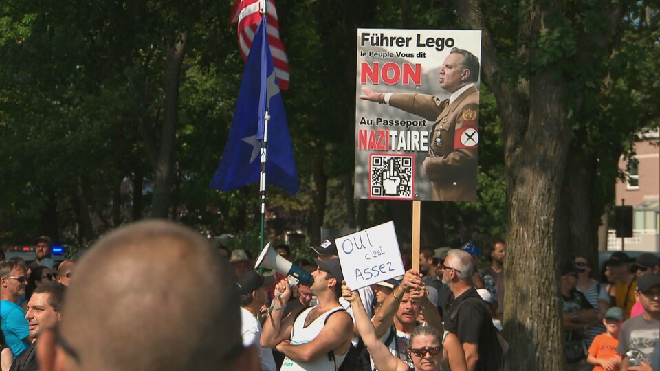 Une pancarte émerge d'une foule et montre François Legault faisant un salut hitlérien dans un uniforme nazi. L'affiche s'oppose au passeport sanitaire.