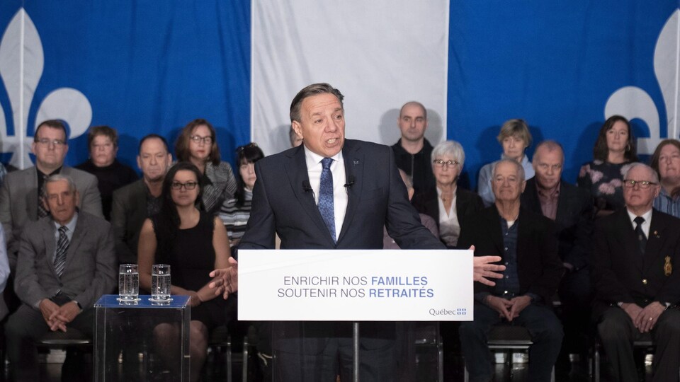 François Legault parle devant un lutrin. Derrière lui, des dizaines de citoyens sont assis devant un grand drapeau du Québec.