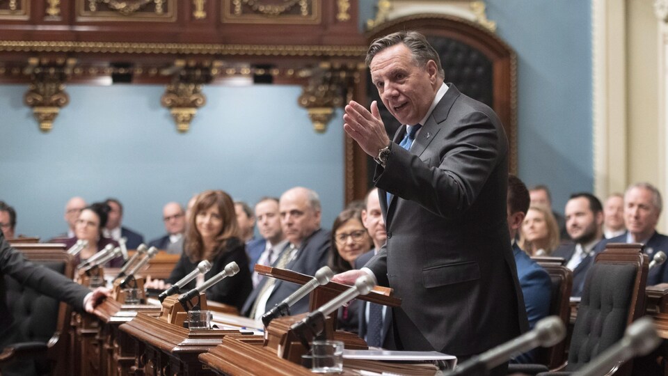 M. Legault lève la main gauche pendant qu'il parle, debout, parmi les députés assis.