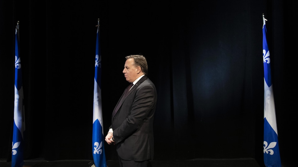 François Legault debout dans une salle de conférence de presse, les bras croisés et les yeux baissés.