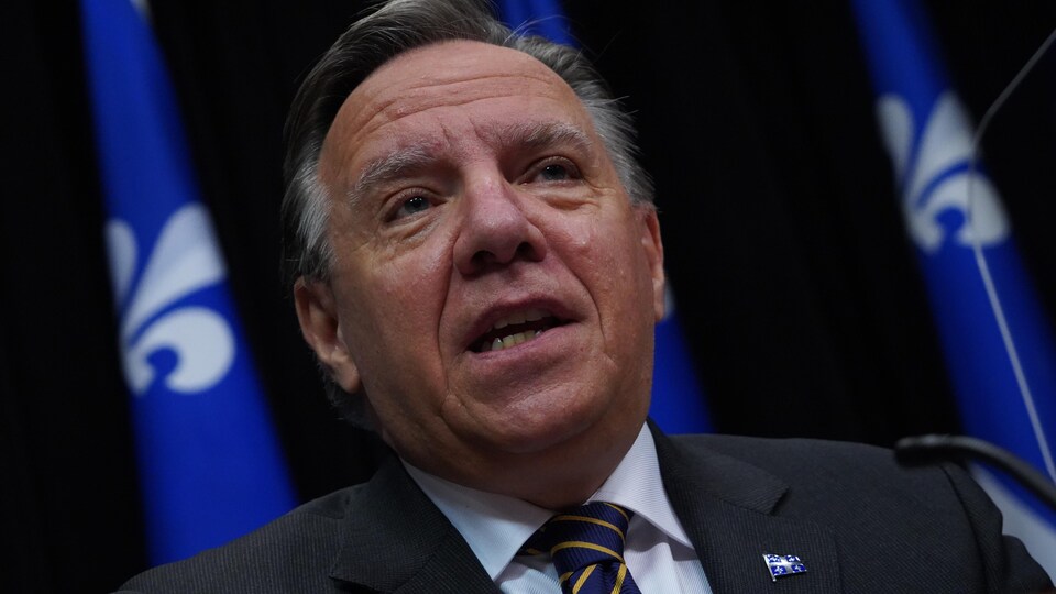 François Legault parle assis à la table de presse devant des drapeaux du Québec. 