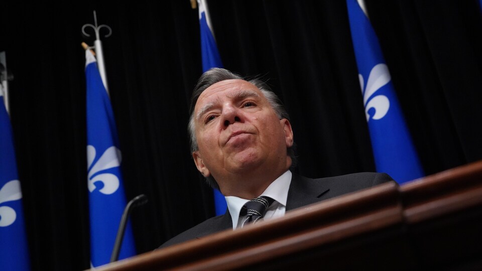 Le premier ministre en complet en conférence de presse, avec des drapeaux du Québec derrière lui