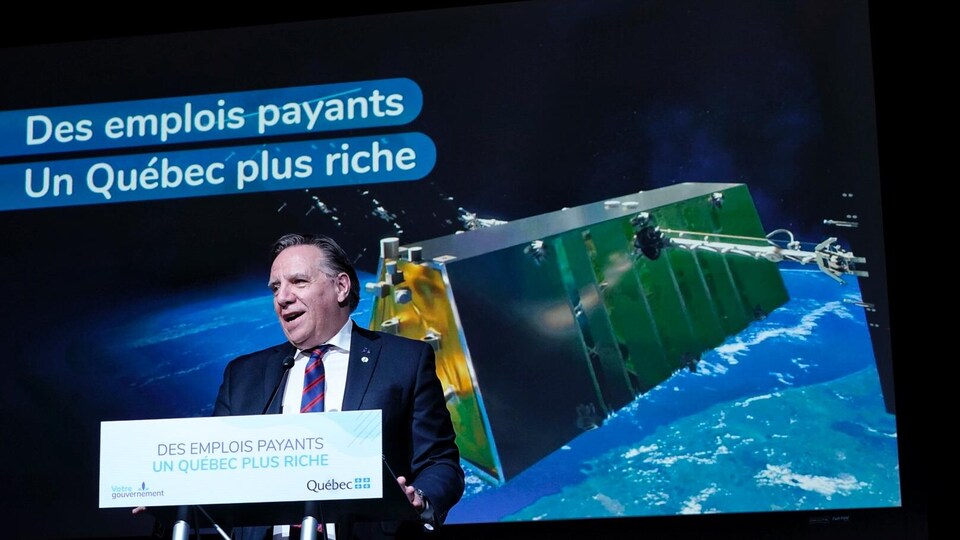François Legault au micro avec, en arrière-plan, l’image d’un satellite dans l'espace.