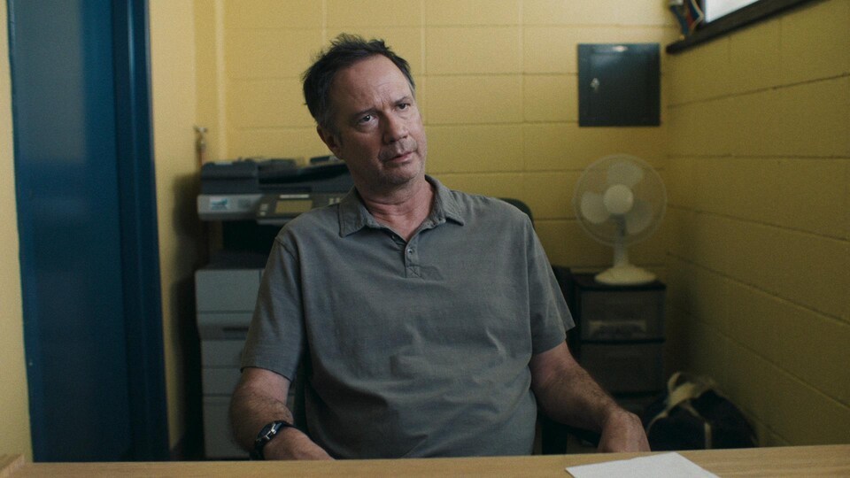 Un homme affiche un air consterné, assis devant un bureau dans une petite pièce. 