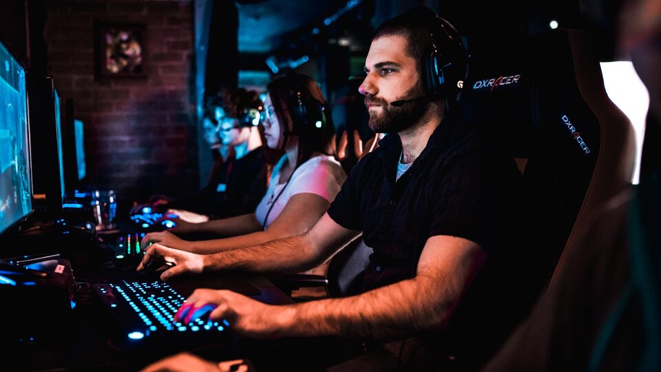 Un homme habillé en noir joue à un jeu vidéo à l'ordinateur à côté d'autres joueurs et joueuses. 