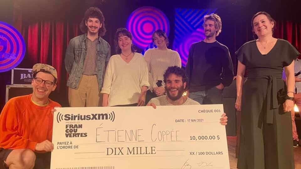 Simon Boulerice et Étienne Coppée, accroupis, tiennent un très grand chèque symbolique sur scène, entourés de plusieurs personnes.