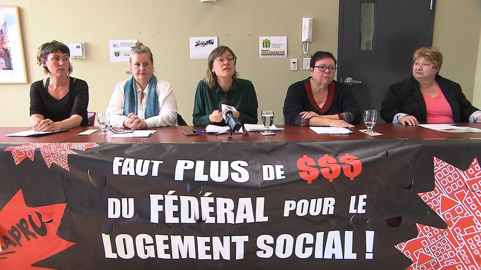 Cinq femmes sont assises devant une table, décorée d'une banderole sur laquelle on peut lire : « Faut plus d'argent du fédéral pour le logement social ».