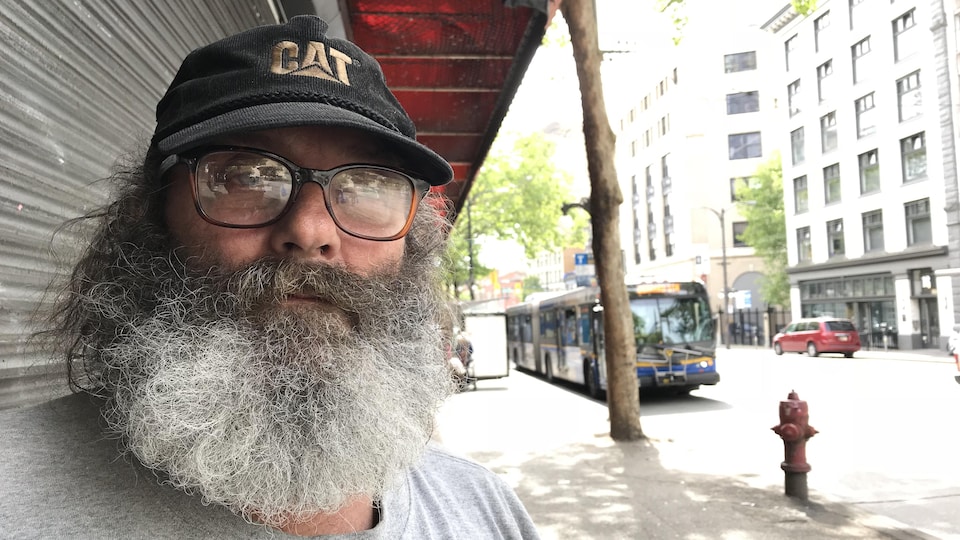 Un homme à la barbe foisonnante poivre et sel arbore une casquette noire et des lunettes à épaisses montures. Il se trouve sur un trottoir du centre-ville.