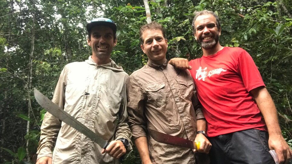 Trois hommes munis de machettes sourient à la caméra.