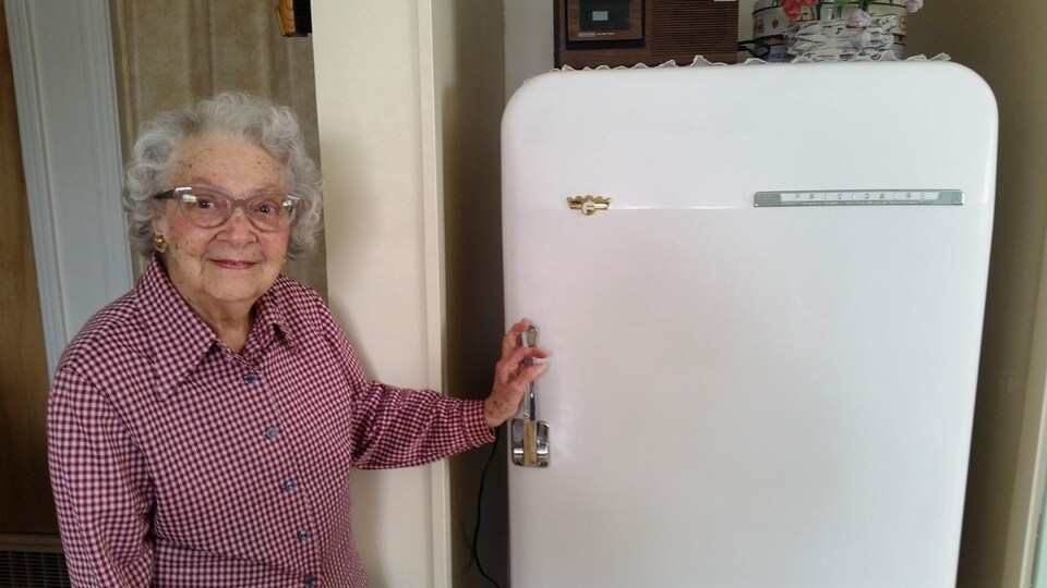 Une dame âgée, coquette, a la main sur la poignée de son vieux frigo qui date de 1953. 