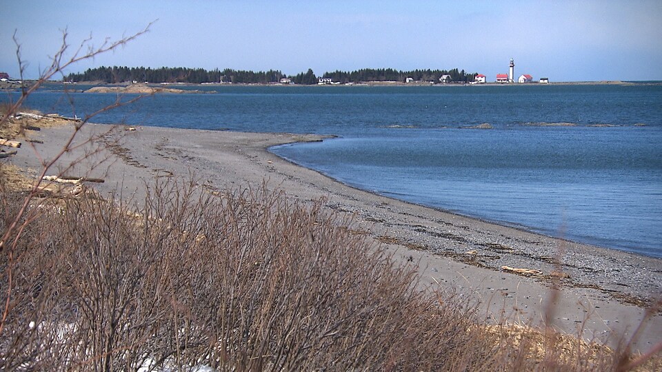 Une plage rencontre le fleuve Saint-Laurent. Au loin, le phare se dresse au bout de la pointe.