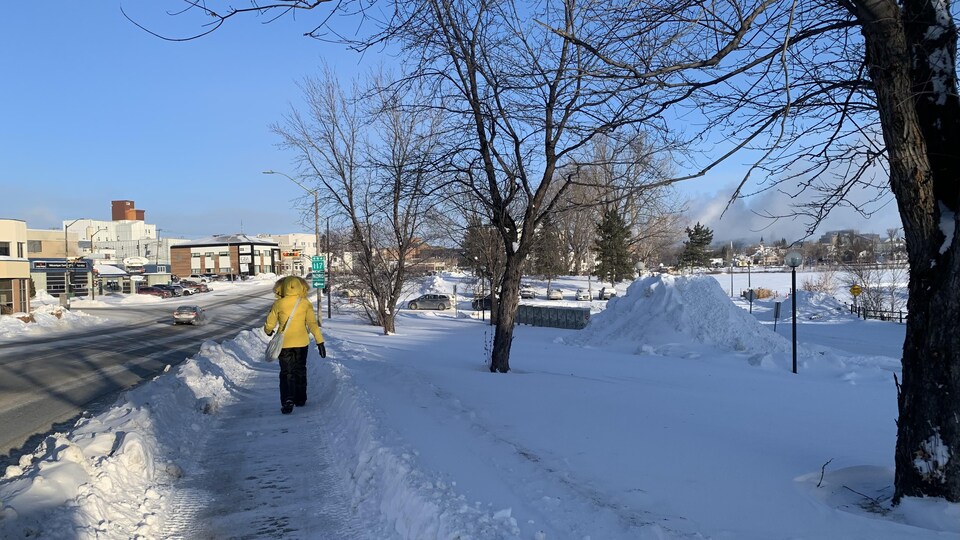 Une femme marche sur un trottoir. Elle porte une capuche, des pantalons de neige et des mitaines pour se protéger du froid.