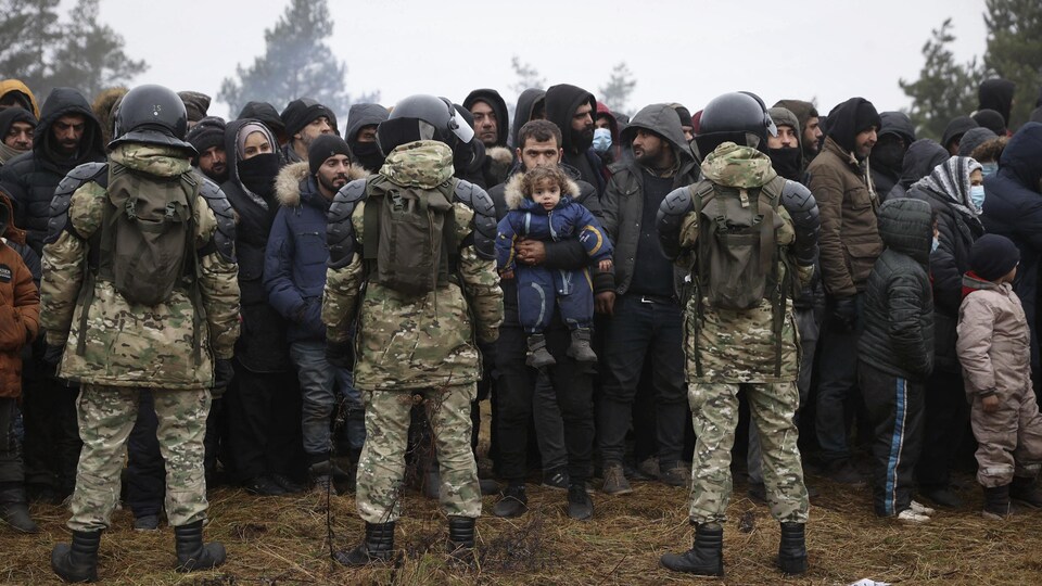 Des gardes vêtus de manteaux aux motifs de camouflage et portant des casques forment une chaîne devant la foule d'hommes, de femmes et d'enfants.