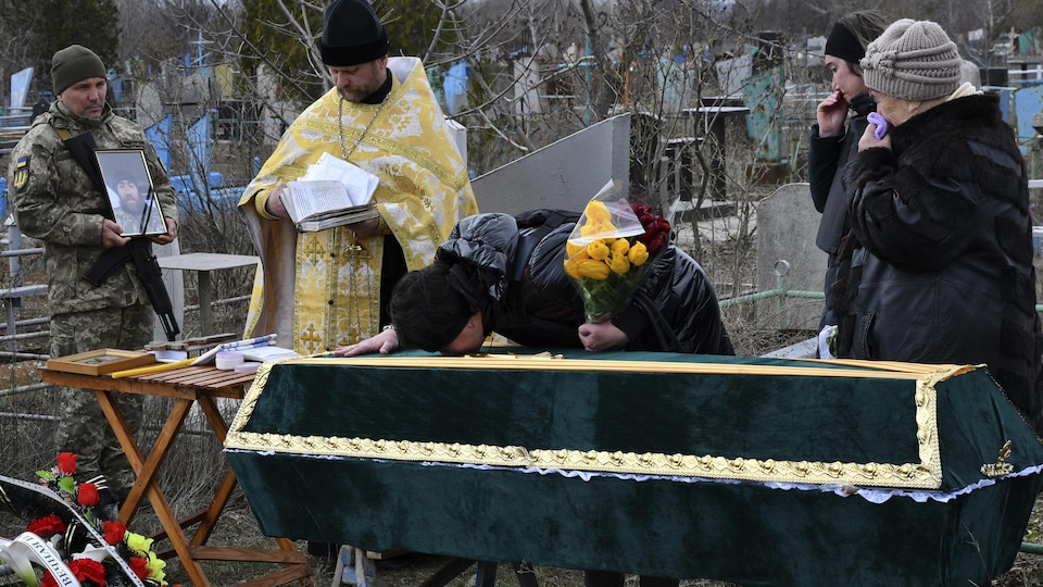 Une personne embrasse un cercueil sous le regard d'un prêtre et d'un militaire qui tient un portrait du défunt. 