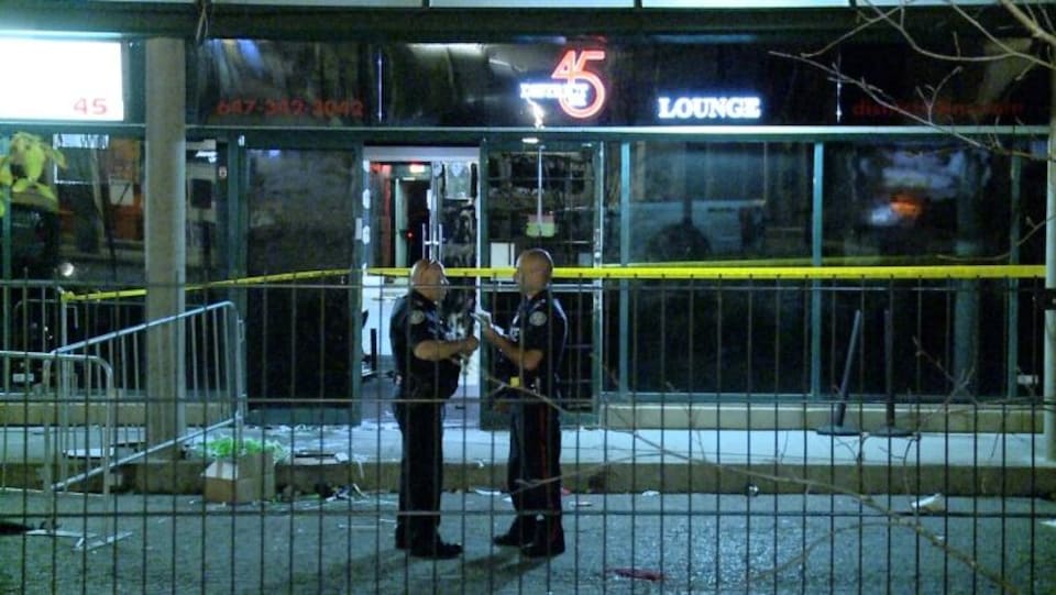 Photo de deux policiers se tenant debout devant la vitrine d'un bar le soir près d'une banderole de police jaune.