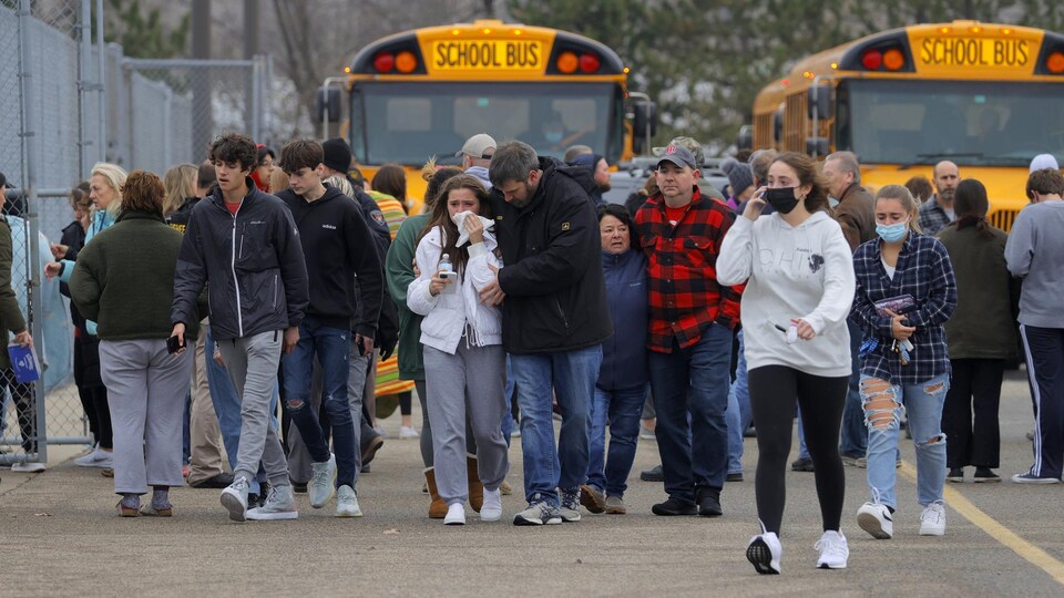 Des élèves et des parents venus chercher leurs enfants marchent dans le stationnement de l'école, où se trouvent deux autobus scolaires.