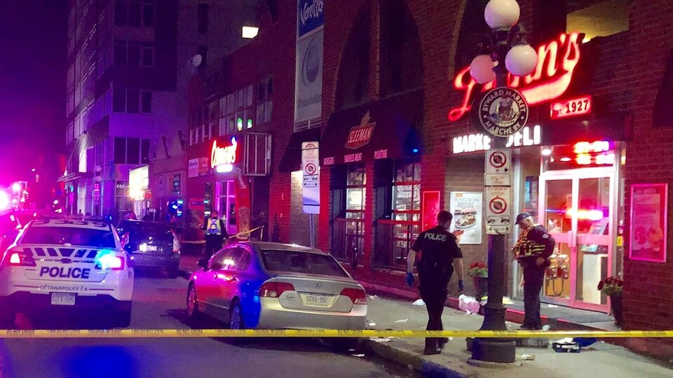 Des véhicules d'urgence sont à l'intérieur d'un périmètre de sécurité, à l'intérieur duquel deux policiers sont en intervention devant un restaurant du marché By.