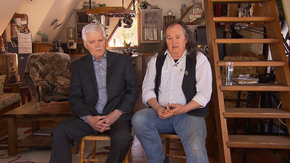 Gaétan Bégin and Pierre Bolduc, sitting in a room of an old house, with a staircase to the right of the image.