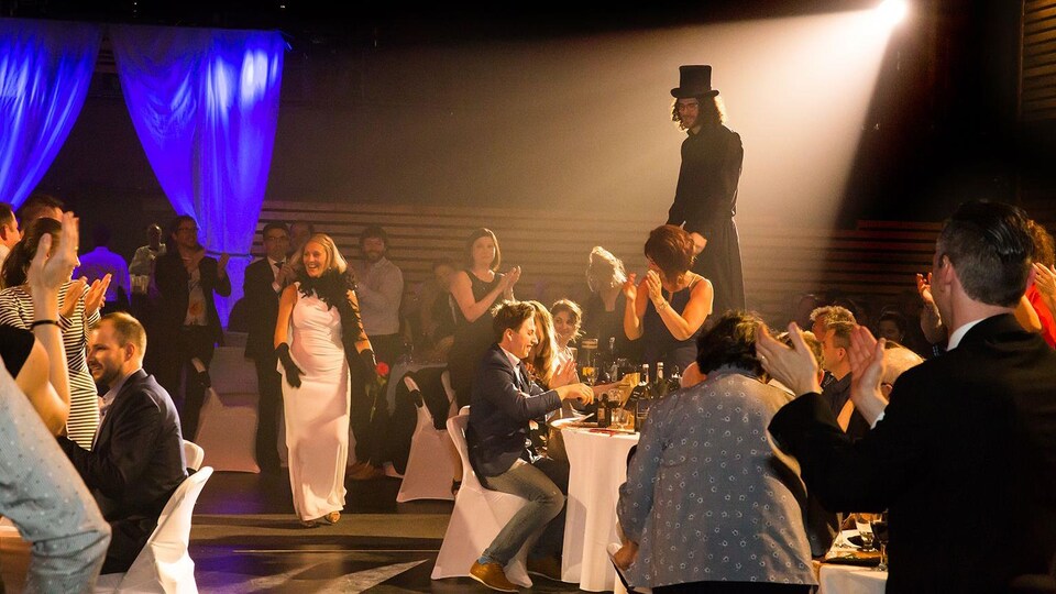 Un homme sur des échasses et une chanteuse se déplacent entre les tables lors d'un gala.