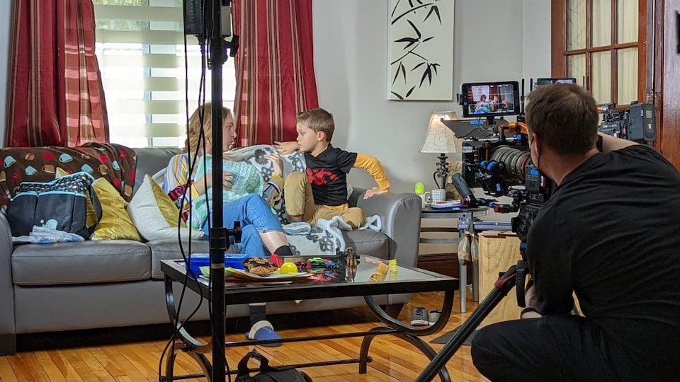 Une femme et un enfant assis sur un sofa sur un plateau de tournage. Un caméraman filme la scène. 
