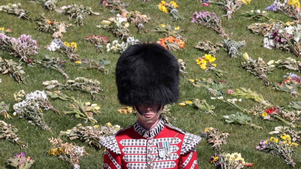 Un homme vêtu d'un habit militaire et d'un immense chapeau poilu se tient bien droit devant un terrain gazonné où ont été déposés des dizaines de bouquets de fleurs.