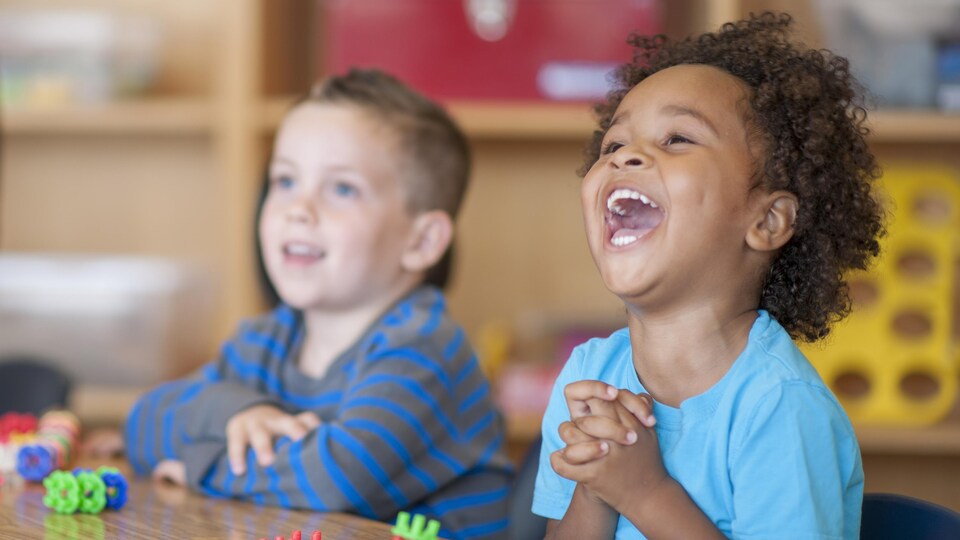 Deux enfants sont assis à une table dans une garderie, dont un qui sourit à pleines dents.
