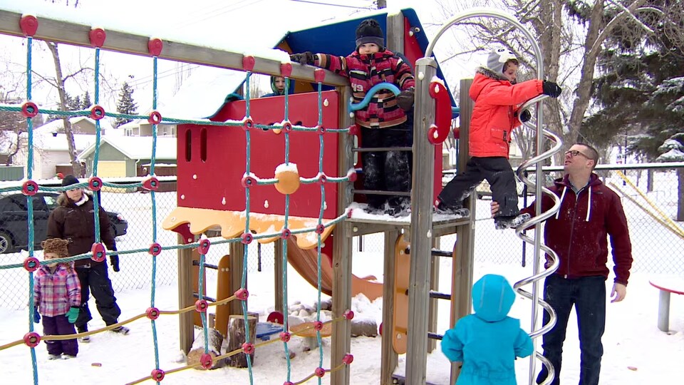 Des enfants jouent dans un module de parc l'hiver sous la surveillance de deux adultes. 
