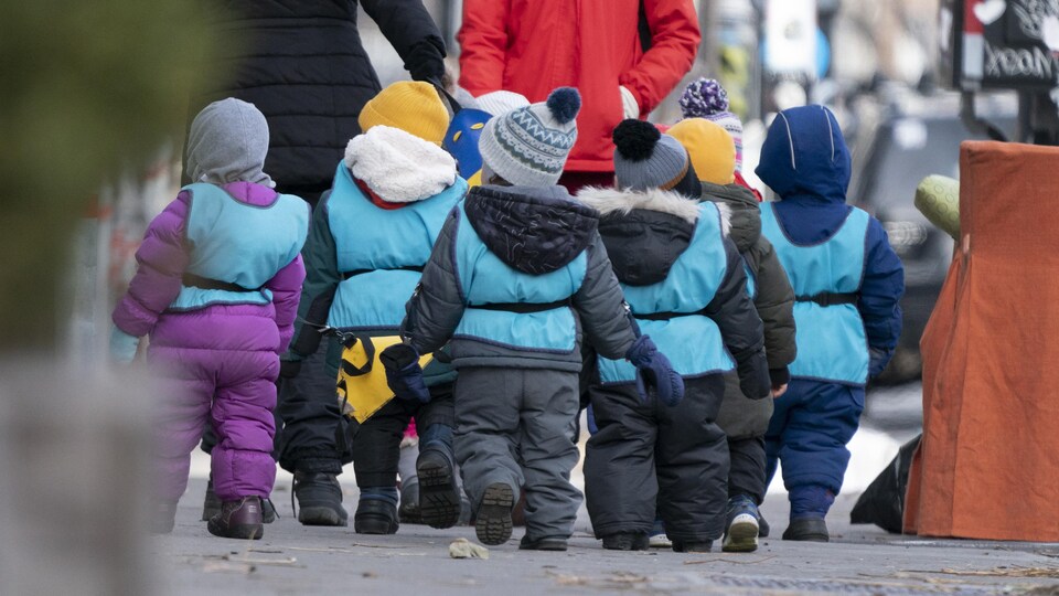 De jeunes enfants dehors et habillés chaudement marchent avec leurs surveillantes. Ils sont de dos.