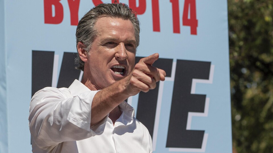 Dehors, Gavin Newsom, devant une affiche référendaire, montre quelque chose du doigt.