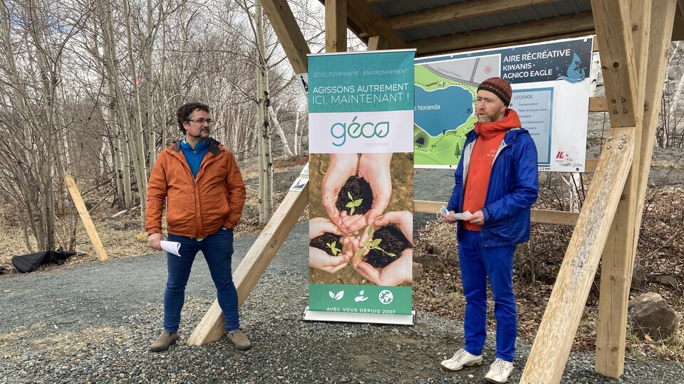 Maurice Duclos et Daniel Richer parlent sous une structure en bois où on a placé une affiche du GÉCO.